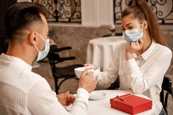 Young couple in medical masks clinking cups of tea near gift box on table in cafe — Fotografia de Stock