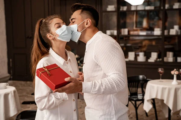 Young man in medical mask holding present while kissing girlfriend in restaurant — Stock Photo