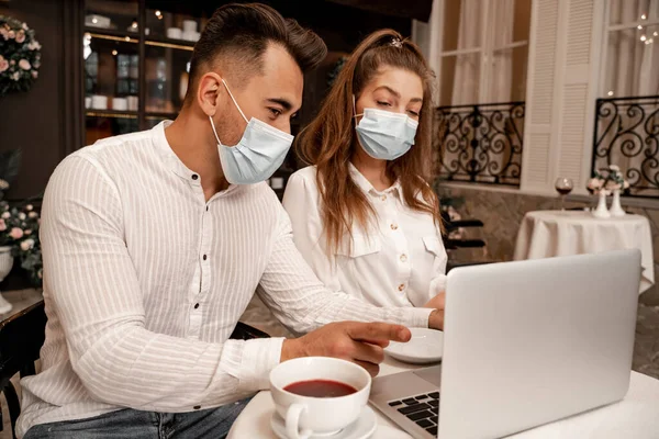 Mann mit medizinischer Maske zeigt mit Finger auf Laptop neben Freundin im Café — Stockfoto