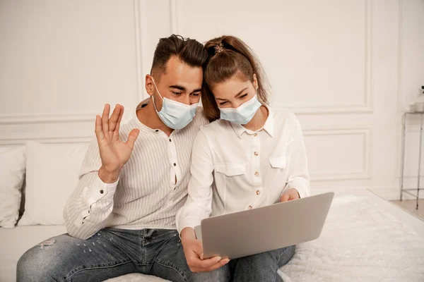 Man in medical mask waving hand during video call near girlfriend at home - foto de stock
