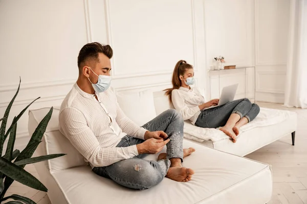 Пара в медицинских масках и джинсах с помощью гаджетов на диване дома — стоковое фото