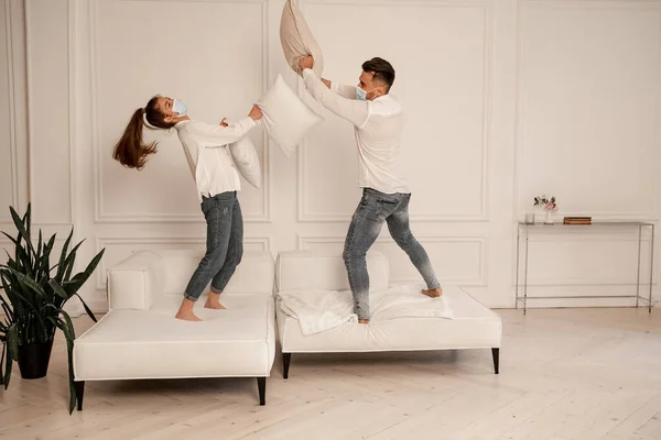 Young couple in medical masks fighting with pillows while having fun at home — Stock Photo