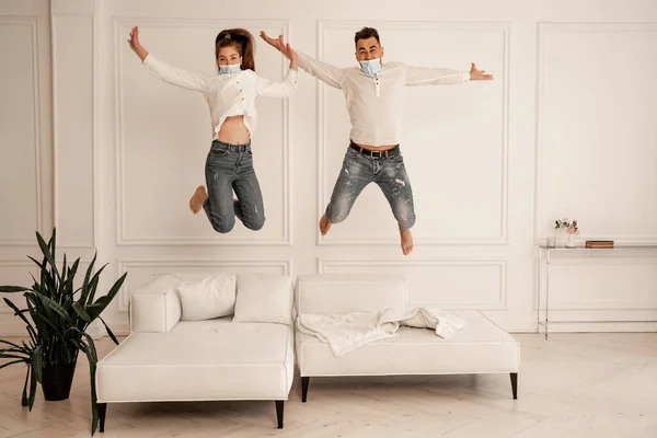Couple in jeans and medical masks having fun while jumping on sofa — Fotografia de Stock