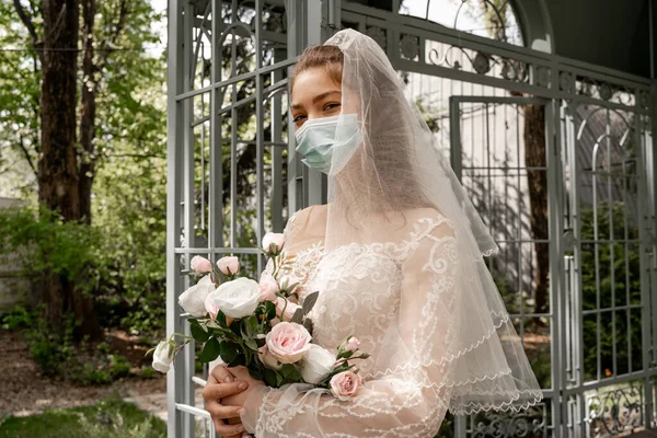 Elegant bride in protective mask looking at camera while holding wedding bouquet outdoors — Photo de stock