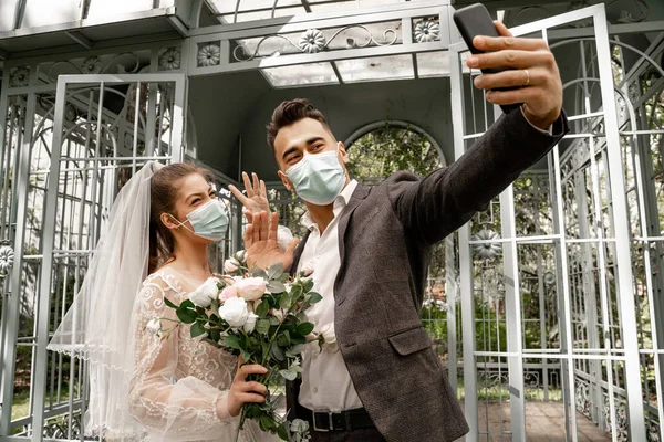 Newlyweds in medical masks taking selfie on mobile phone in park — Photo de stock