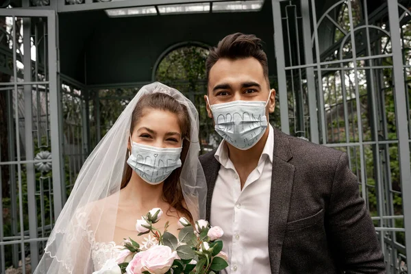 Newlyweds in medical masks with bride and groom lettering looking at camera outdoors — Photo de stock