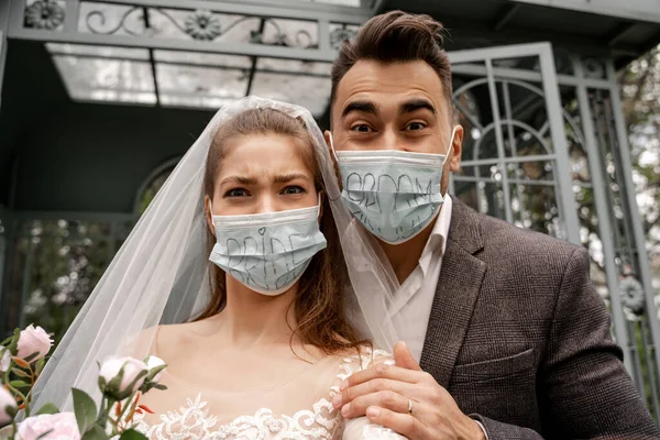 Excited newlyweds in safety masks with bride and groom lettering looking at camera in park — Stock Photo