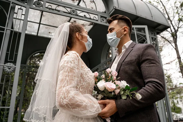 Low angle view of newlyweds in protective masks looking at each other outdoors — Stock Photo