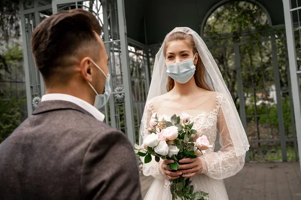 Young bride in medical mask holding wedding bouquet near groom on blurred foreground - foto de stock