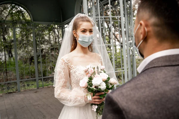 Elegant bride in medical mask holding wedding bouquet near blurred groom in park - foto de stock