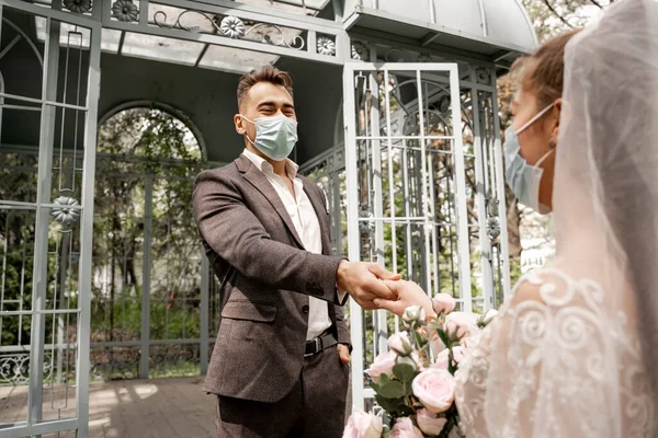 Young newlyweds in safety masks holding hands in park on blurred foreground — Foto stock