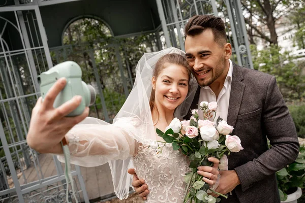 Cheerful bride taking selfie with groom on digital camera in park, blurred foreground — Fotografia de Stock
