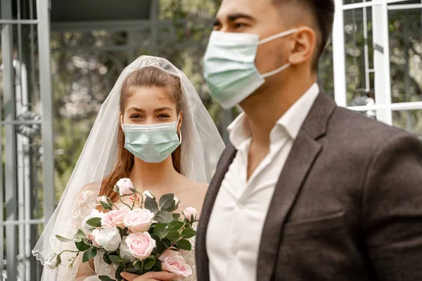 Молода жінка з весільним букетом дивиться на камеру біля розмитого нареченого в медичній масці — стокове фото