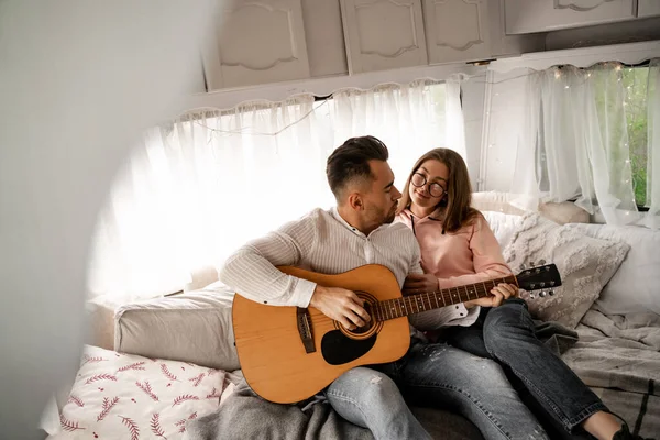 Young man playing guitar while resting with girlfriend in camper on blurred foreground - foto de stock