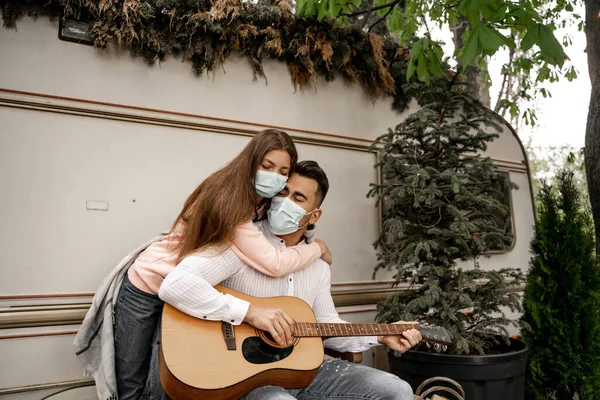 Молода жінка в медичній масці обіймає хлопця, який грає на гітарі біля кемпера — стокове фото