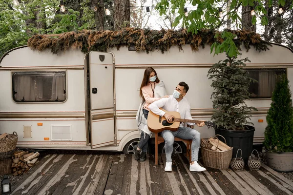 Людина в медичній масці грає на гітарі біля дівчини і трейлері в кемпінгу — стокове фото