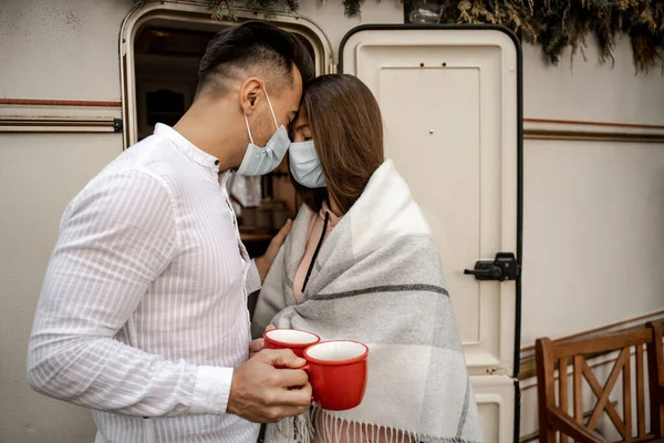 Young couple in medical masks standing with cups of coffee and closed eyes near camper - foto de stock