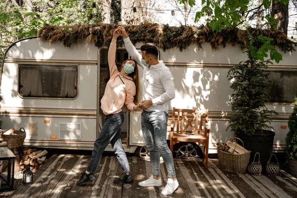 Young couple in protective masks dancing near camper in forest — Stock Photo