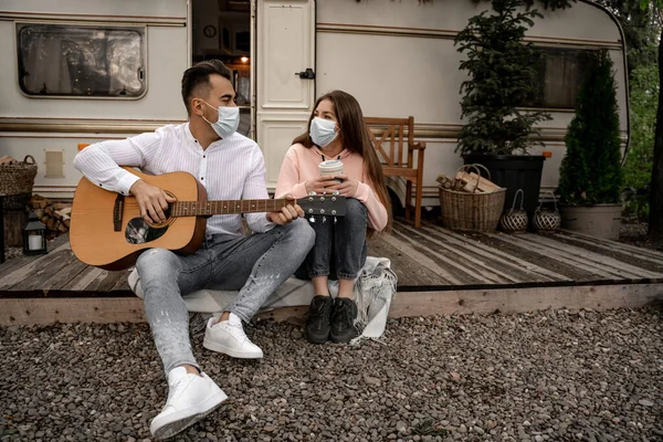 Чоловік в медичній масці грає на гітарі біля жінки з термо чашкою в кемпінгу — стокове фото