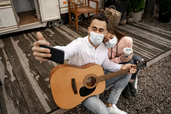 Чоловік з гітарою бере селфі з дівчиною в медичній масці на відкритому повітрі в кемпінгу — стокове фото