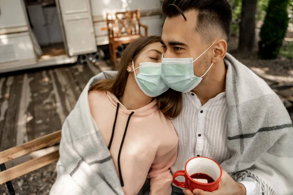 Young man in medical mask holding cup of coffee while sitting with girlfriend outdoors - foto de stock