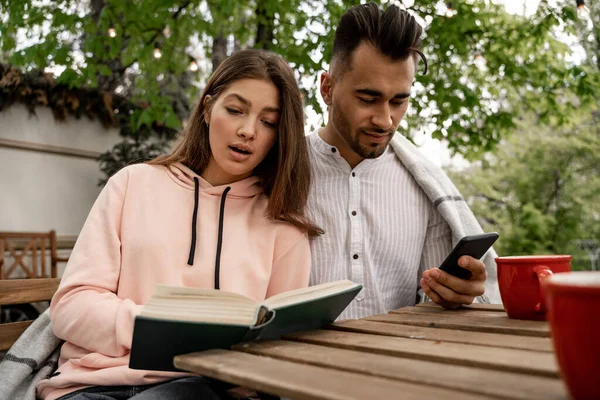 Pretty woman reading book near man chatting on smartphone outdoors - foto de stock