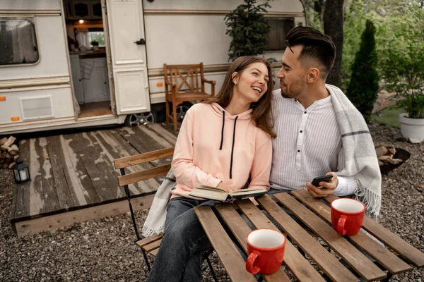 Young woman with book laughing near man with smartphone near camper — Stock Photo