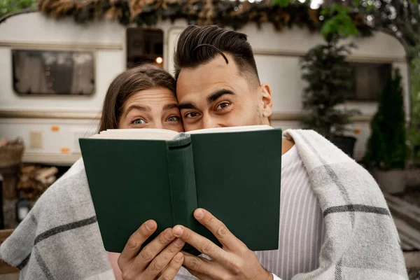 Cheerful couple obscuring faces with book while resting near trailer on blurred background - foto de stock