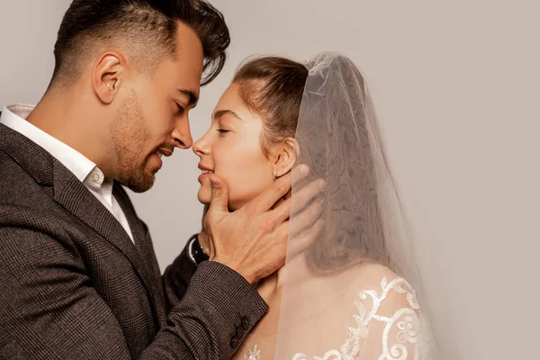 Seitenansicht des jungen Mannes berühren Gesicht der charmanten Braut isoliert auf grau — Stockfoto