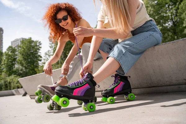 Mulher encaracolado em óculos de sol amarrando atacadores no patins perto amigo no parque — Fotografia de Stock