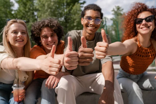 Amigos multiétnicos borrosas sonriendo y mostrando los pulgares hacia arriba en el parque de la ciudad - foto de stock