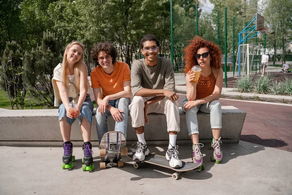 Mujeres jóvenes felices con copas de plástico sentadas en la frontera junto con patinadores interracial - foto de stock