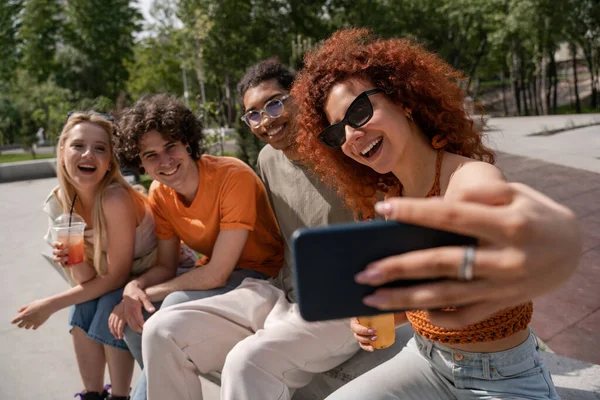 Mujer rizada y alegre en gafas de sol tomando selfie con amigos multiétnicos - foto de stock