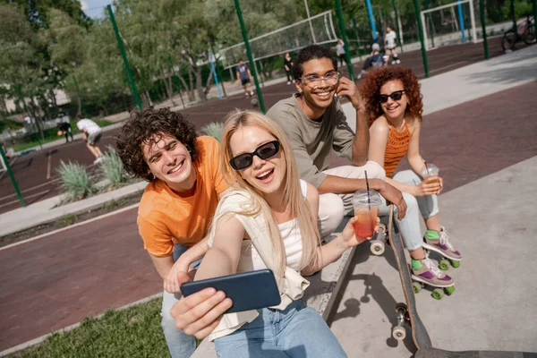 Happy young woman in sunglasses taking selfie with multiethnic skaters outdoors — Stock Photo