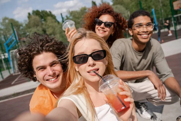 Giovane donna in occhiali da sole bere cocktail rinfrescante vicino amici multietnici felici — Foto stock