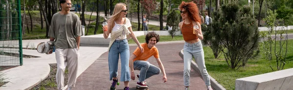 Happy multiethnic friends skating in park with cocktails, banner — Stock Photo