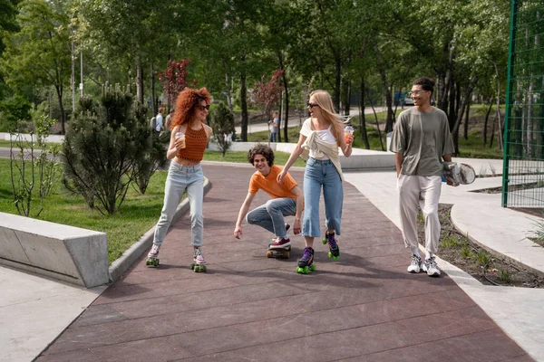 Sorridente uomo africano americano a piedi vicino amici felici pattinaggio nel parco — Foto stock