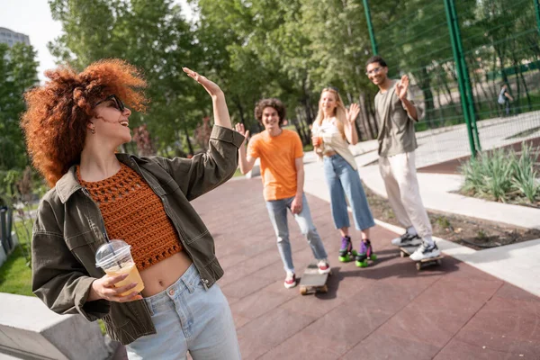 Young excited woman with refreshing drink waving hand to blurred multiethnic skaters — Stock Photo