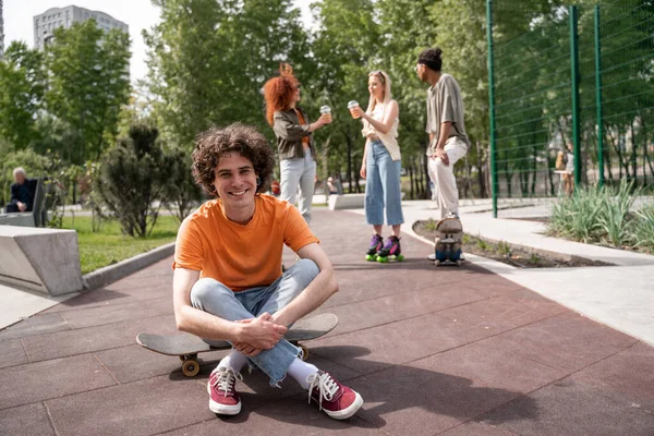 Smiling curly man sitting on skateboard with crossed legs near blurred multiethnic friends on background — Stock Photo