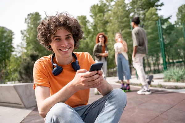 Rizado hombre con el teléfono móvil sonriendo a la cámara cerca borrosa patinadores multiétnicos - foto de stock