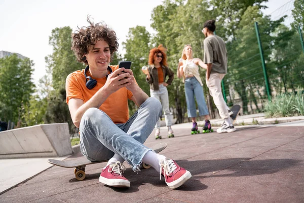 Junger lächelnder Mann sitzt auf Skateboard und sendet Nachrichten auf dem Smartphone neben verschwommenen interrassischen Skatern — Stockfoto