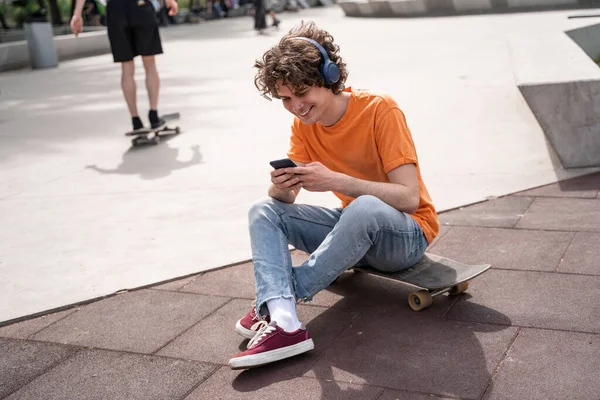 Happy man in headphones sitting on skateboard and using smartphone in skate park — Stock Photo