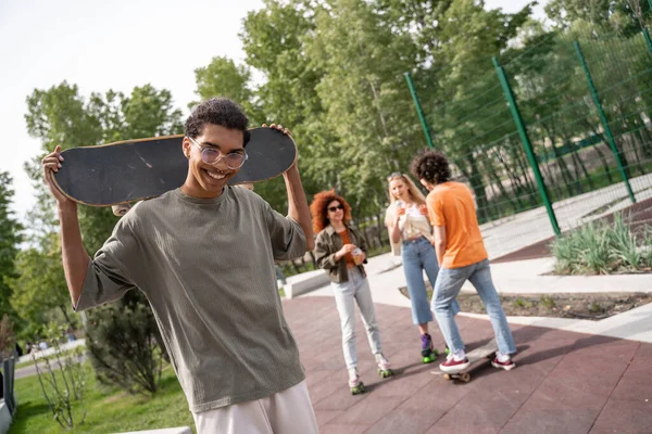 Hombre afroamericano feliz con monopatín mirando a la cámara cerca de amigos multiétnicos - foto de stock