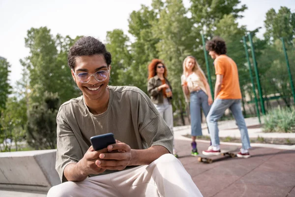 African american man in eyeglasses messaging on cellphone near skaters on blurred background — Stock Photo