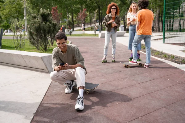 Alegre africano amerciano hombre charlando en el teléfono móvil cerca de amigos en skate park - foto de stock