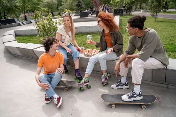 Alegres patinadores multiétnicos hablando cerca de sabrosa pizza en skate park - foto de stock