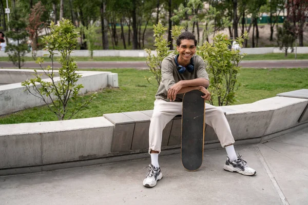 Fröhlicher afrikanisch-amerikanischer Mann lächelt in die Kamera, während er auf einer Bank mit Skateboard sitzt — Stockfoto