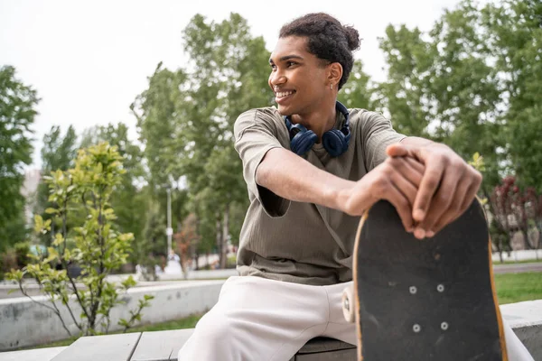Young and joyful african american skater looking away while sitting in skate park — Stock Photo
