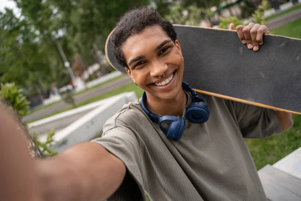 Piercing afro-americano homem com fones de ouvido e skate sorrindo para a câmera — Fotografia de Stock