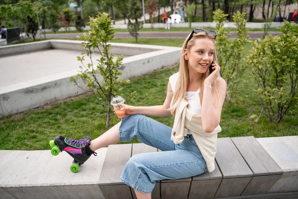 Smiling roller skater with cocktail sitting on border bench and talking on mobile phone — Stock Photo